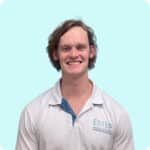 A young man with wavy brown hair smiles at the camera. He is wearing a white collared shirt with a light blue accent and EMED written on the left side. The background is a solid pale blue.
