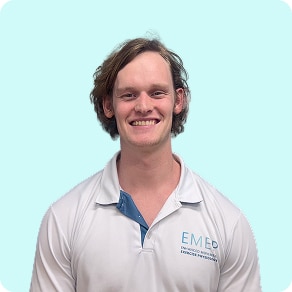 A young man with wavy brown hair smiles at the camera. He is wearing a white collared shirt with a light blue accent and EMED written on the left side. The background is a solid pale blue.