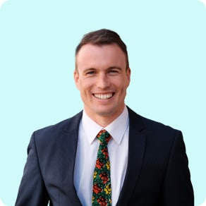A man in a dark suit, white shirt, and a colourful floral tie smiles at the camera in front of a solid light blue background.