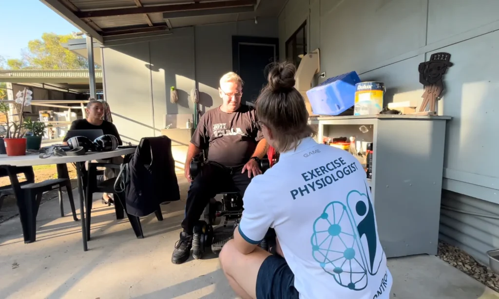 A person in a wheelchair sits on a verandah facing a woman wearing an Exercise Physiologist shirt; two other people are seated at a table nearby, and various items are on a shelf in the background.