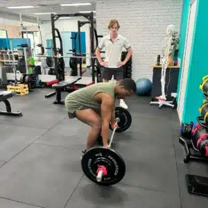 A person in a gym prepares to lift a barbell while another person stands behind, observing. Gym equipment, mirrors, and a skeleton model are visible in the background.
