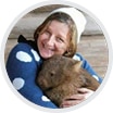 A person wearing a blue jumper with white polka dots and a white beanie smiles whilst hugging a wombat.