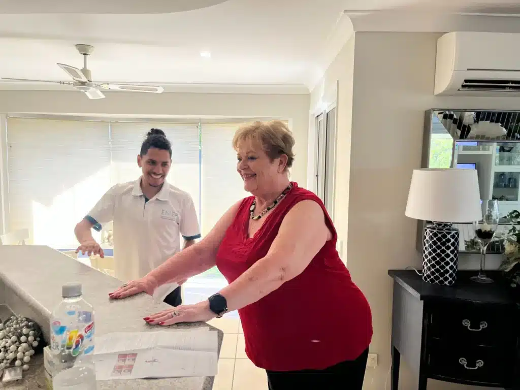 A smiling woman in a red top leans on a kitchen worktop while a man in a white shirt stands nearby, both appearing cheerful in a bright, modern home. Objects like a water bottle and papers sit on the worktop.