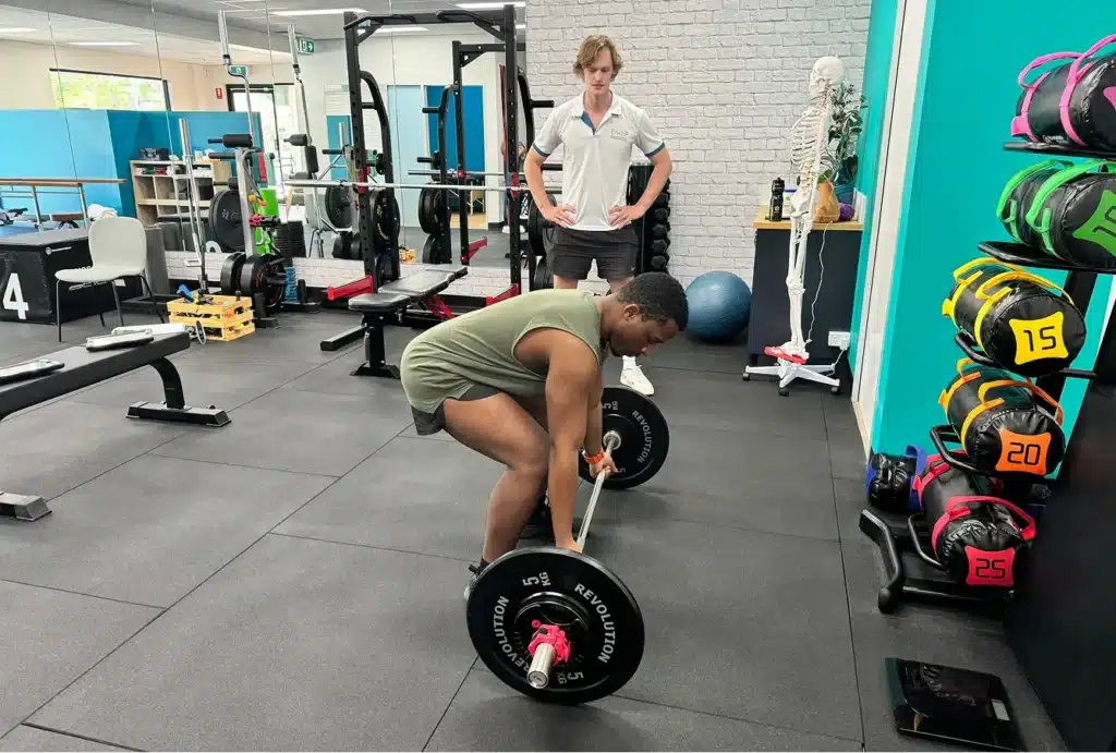 A person in a green shirt prepares to deadlift a barbell in a gym, whilst another person stands behind, observing and giving guidance. Weight plates and gym equipment are visible in the background.