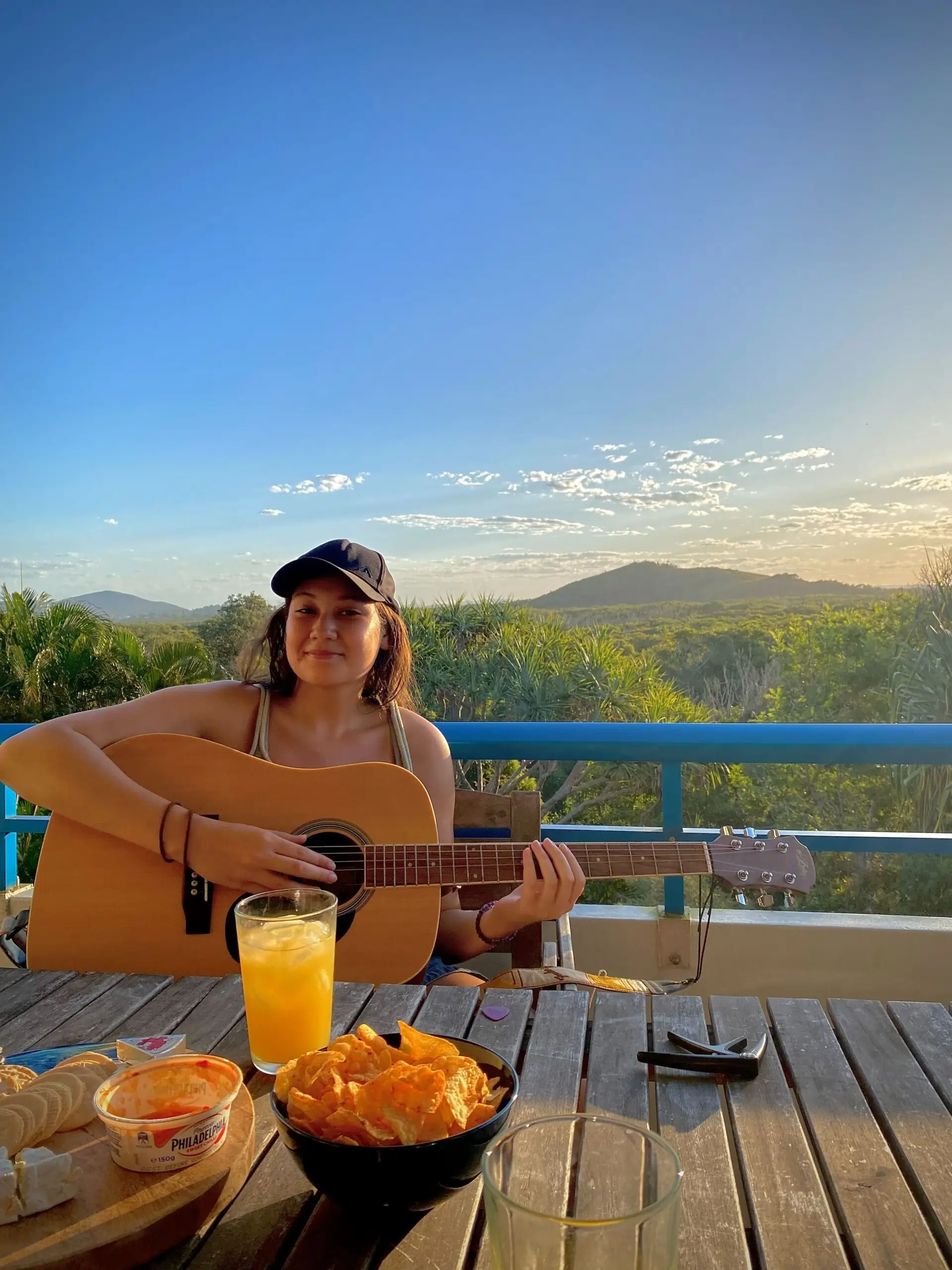 A person wearing a cap plays an acoustic guitar on an outdoor patio at sunset, with snacks, a drink, and a scenic view of green hills and a clear sky in the background.