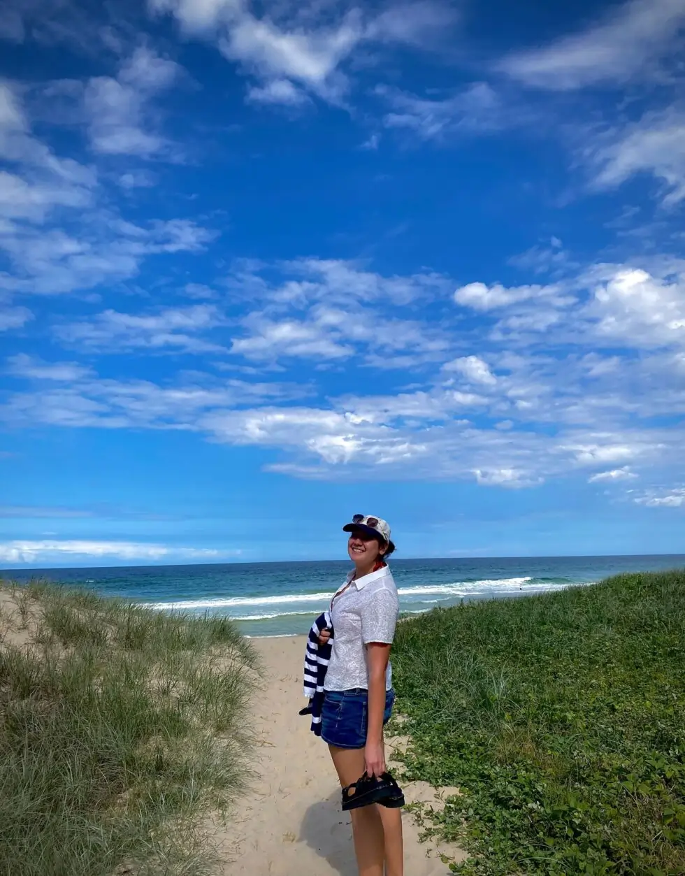 A person in shorts and a hat stands smiling on a sandy path between grassy dunes, holding sandals, with the sea and a blue sky filled with clouds in the background.