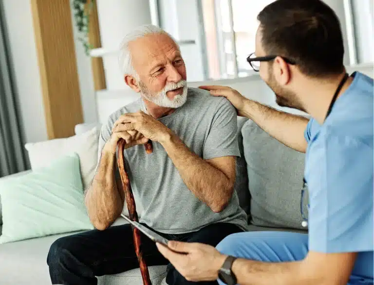 An elderly man with a walking stick sits on a sofa, smiling and talking to a healthcare professional in scrubs, who is holding a clipboard and gently touching the man's shoulder.