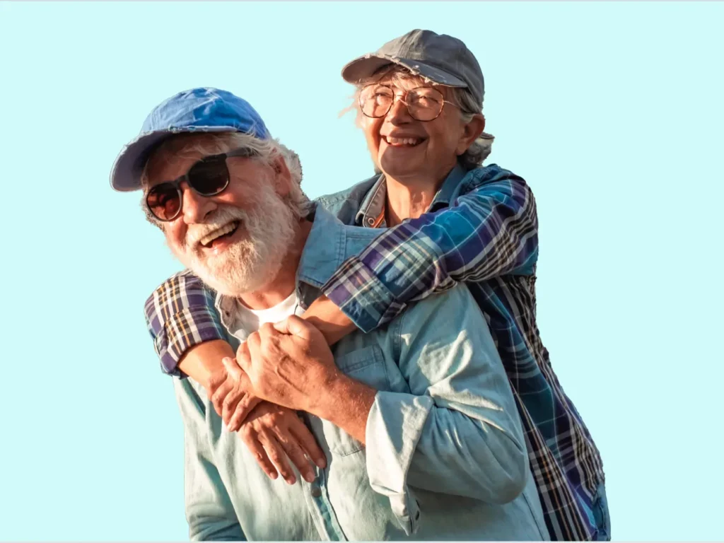 An elderly woman in a cap and glasses gives a piggyback ride to an elderly man wearing sunglasses and a blue cap. Both are smiling happily against a light blue background.