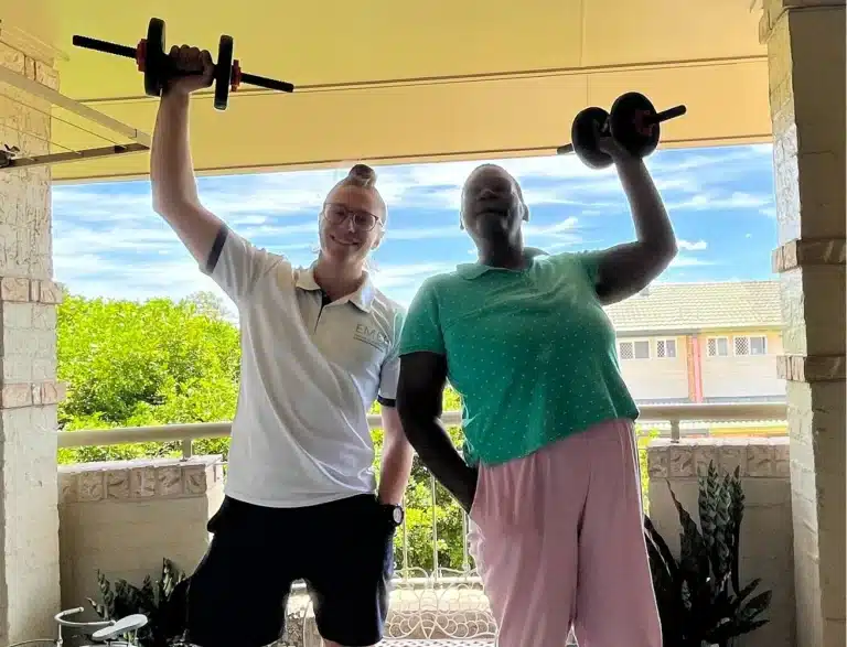 Two people stand on a balcony, both smiling and lifting dumbbells above their heads. One wears a white shirt and black shorts, the other wears a green shirt and pink trousers. There are plants and a building in the background.