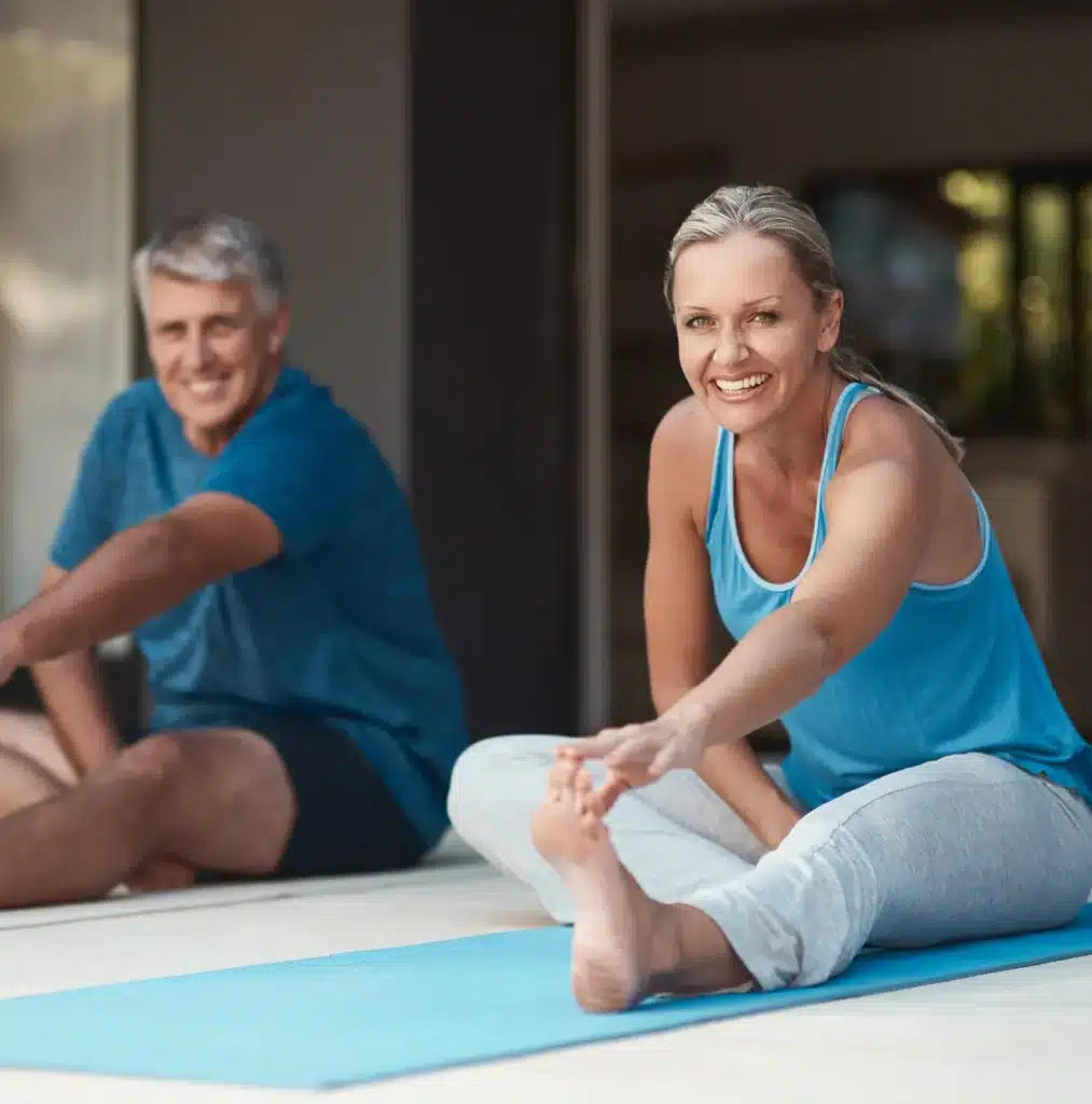 A smiling woman and man sit on yoga mats indoors, stretching their legs and reaching for their toes during a fitness or yoga session.
