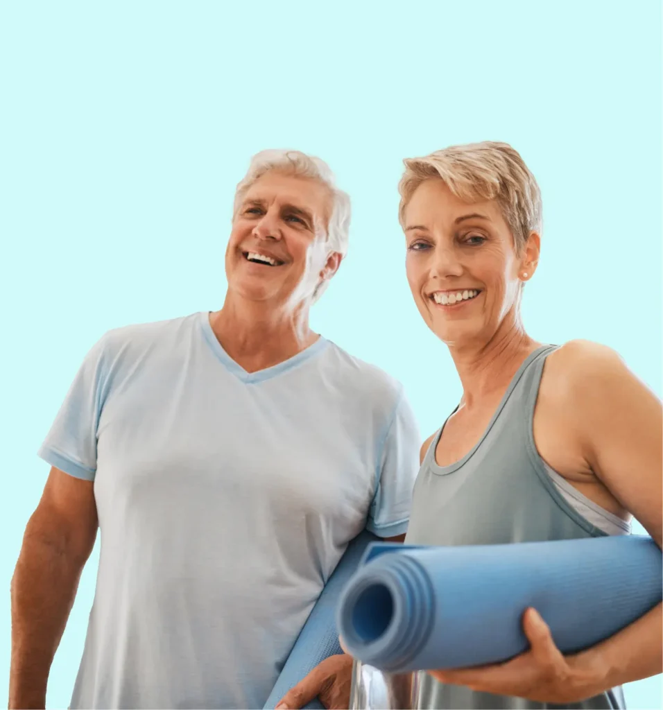 A smiling older man and woman stand together, each holding a rolled-up yoga mat, dressed in light athletic clothes against a light blue background.