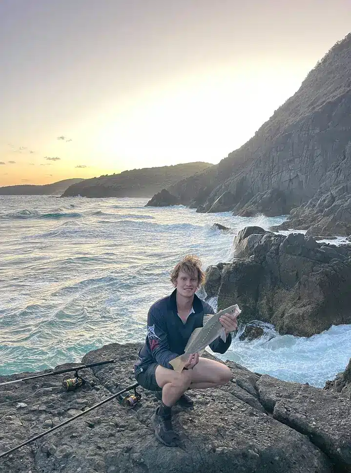A person kneels on a rocky shore at sunset, holding a large fish. Waves crash against the rocks nearby, with a fishing rod lying on the ground. Hills rise in the background under a clear sky.