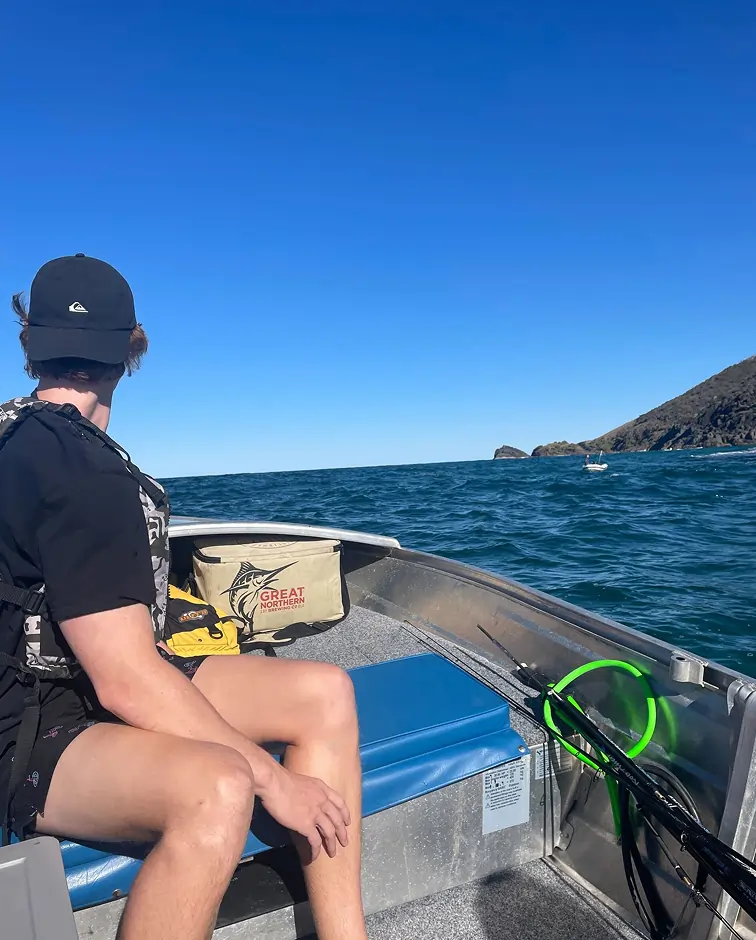 A person wearing a black shirt, cap, and life jacket sits on a boat looking out at the sea under a clear blue sky, with a rocky coastline visible in the distance and fishing gear beside them.