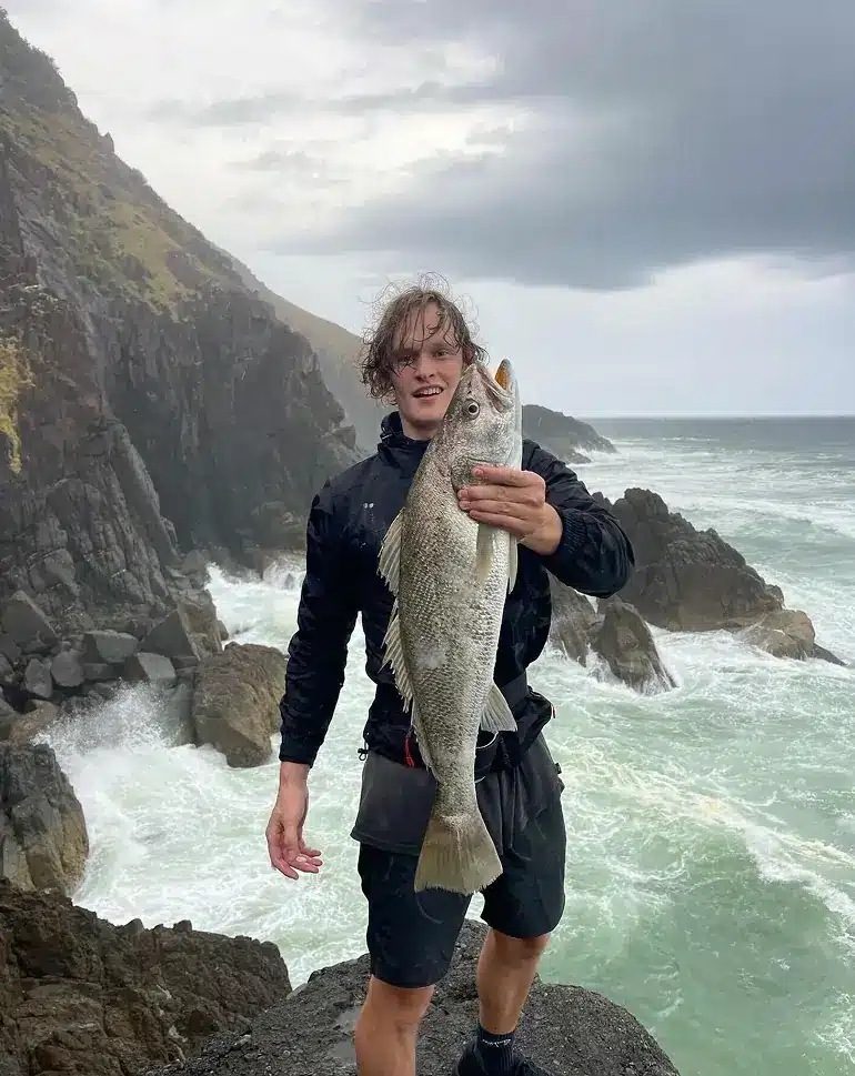 A person standing on coastal rocks holds a large fish, with rough waves crashing behind them and steep cliffs under a cloudy sky.