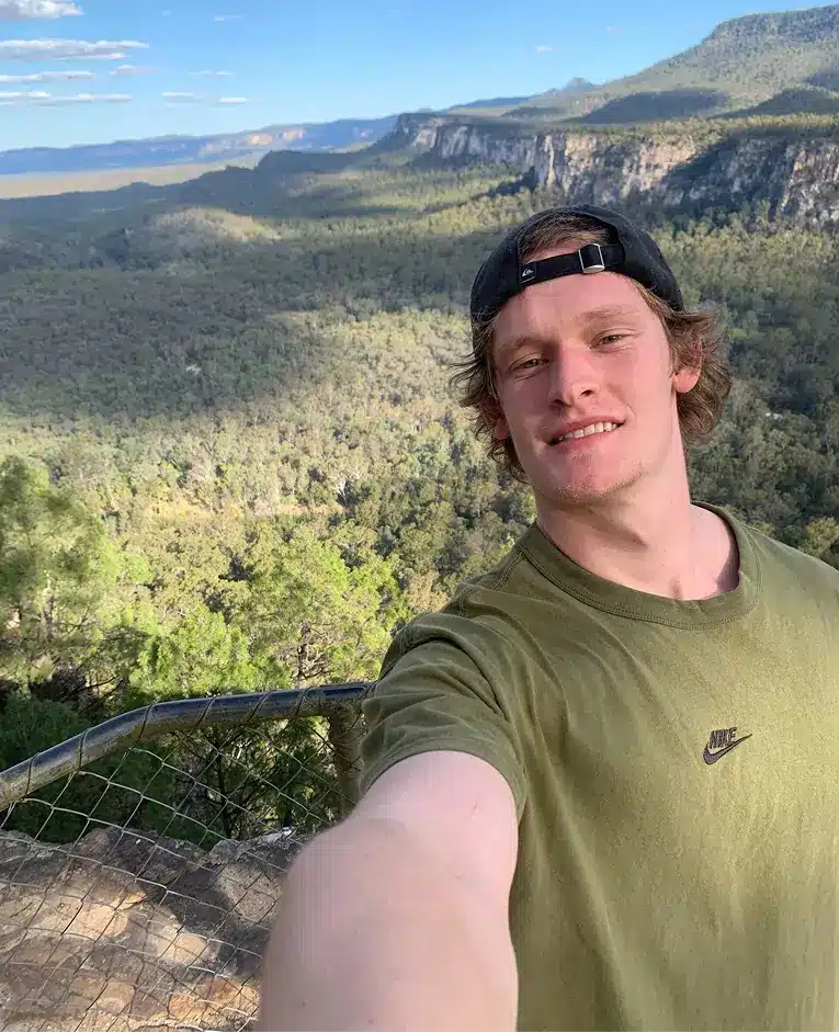 A young man in a green Nike shirt and backwards cap takes a selfie on a lookout, with a vast forest, cliffs, and mountains stretching into the distance under a blue sky.