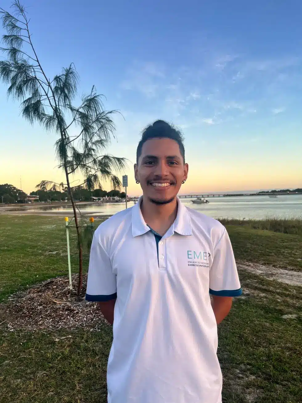 A man with short curly hair, a moustache, and a goatee smiles while standing outdoors near a body of water, wearing a white EMB logo polo shirt. A tree and grassy area are in the background under a blue sky.