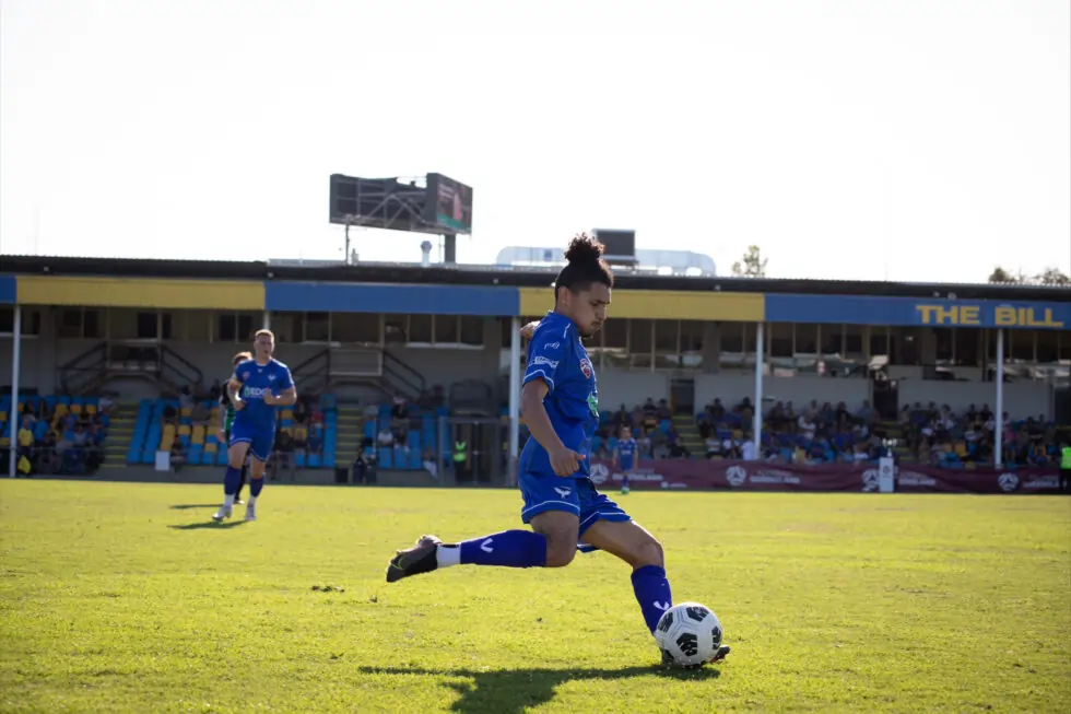 A footballer in a blue kit prepares to kick a ball on a grassy pitch, with spectators sitting in stands and another player in the background.