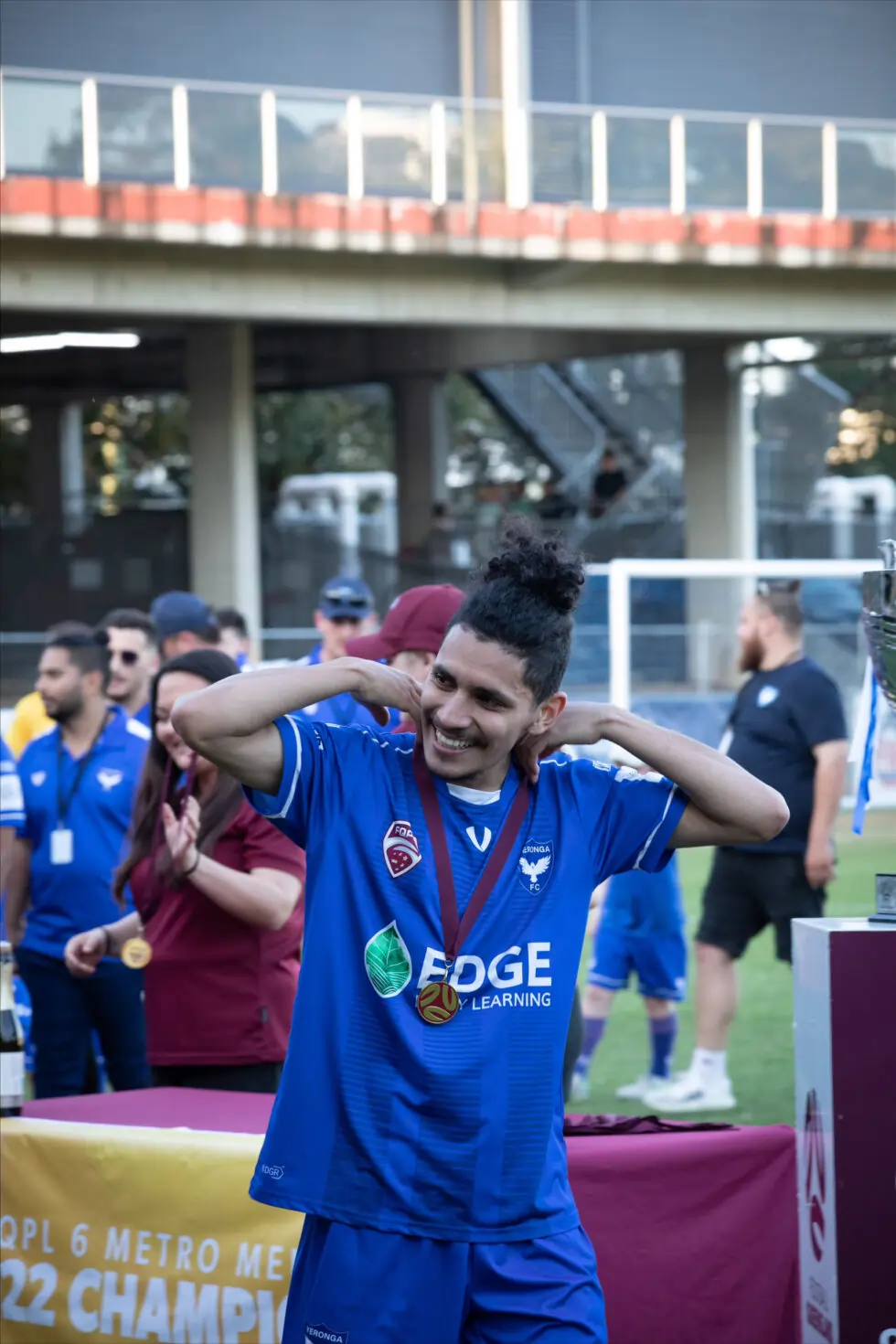 A smiling footballer in a blue kit celebrates on a pitch, wearing a medal around his neck. People and a trophy are visible in the background, indicating a championship event.
