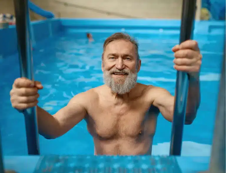 An older man with a grey beard smiles while holding onto the metal ladder of a swimming pool, appearing to climb out of the water. The indoor pool is bright blue, and another swimmer is visible in the background.