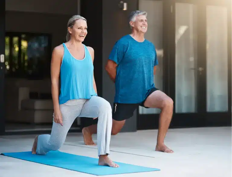 A smiling older woman and man do lunges together; she is on a yoga mat wearing a blue vest top and grey trousers, he is standing behind her in a blue shirt and shorts. They appear to be exercising outdoors.