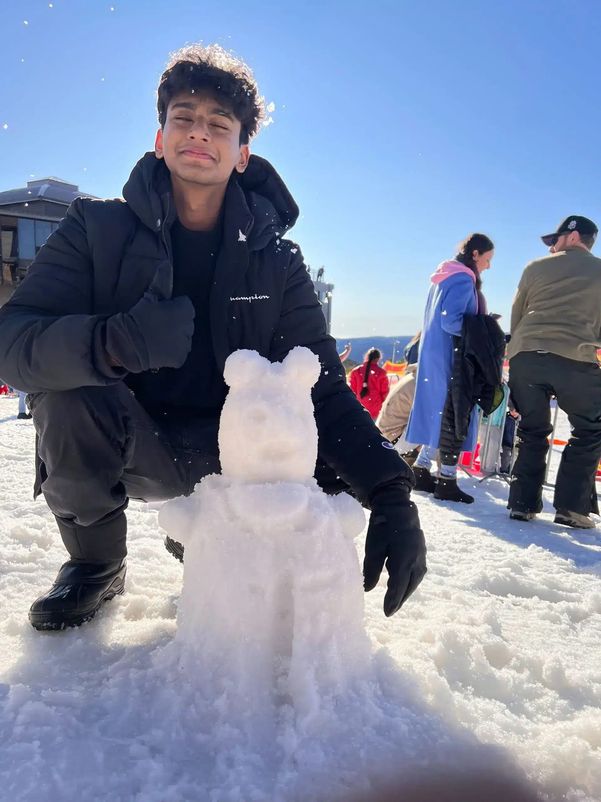 A young person in a black jacket and gloves smiles while kneeling next to a snow sculpture shaped like a bear. Other people and a building are visible in the snowy background under clear blue sky.