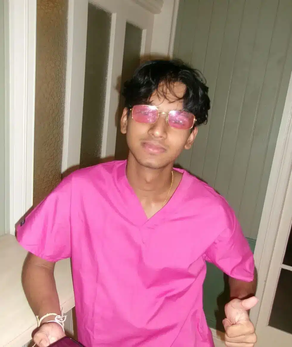 A young man wearing pink scrubs and pink-tinted glasses stands indoors, giving a thumbs up with one hand and holding a mobile in the other, with a slight smile on his face.