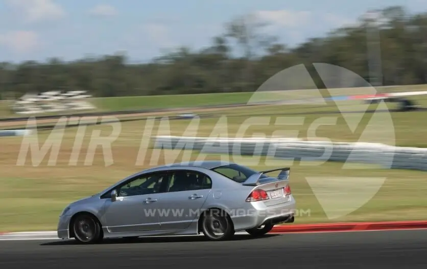A silver saloon with a rear spoiler is speeding around a racetrack, leaning slightly as it takes a corner. The background is blurred, showing grassy areas and trees, emphasising the car’s motion.