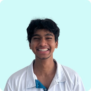 A young man with dark hair smiles broadly, wearing a white collared shirt over a blue shirt, standing against a light blue background.