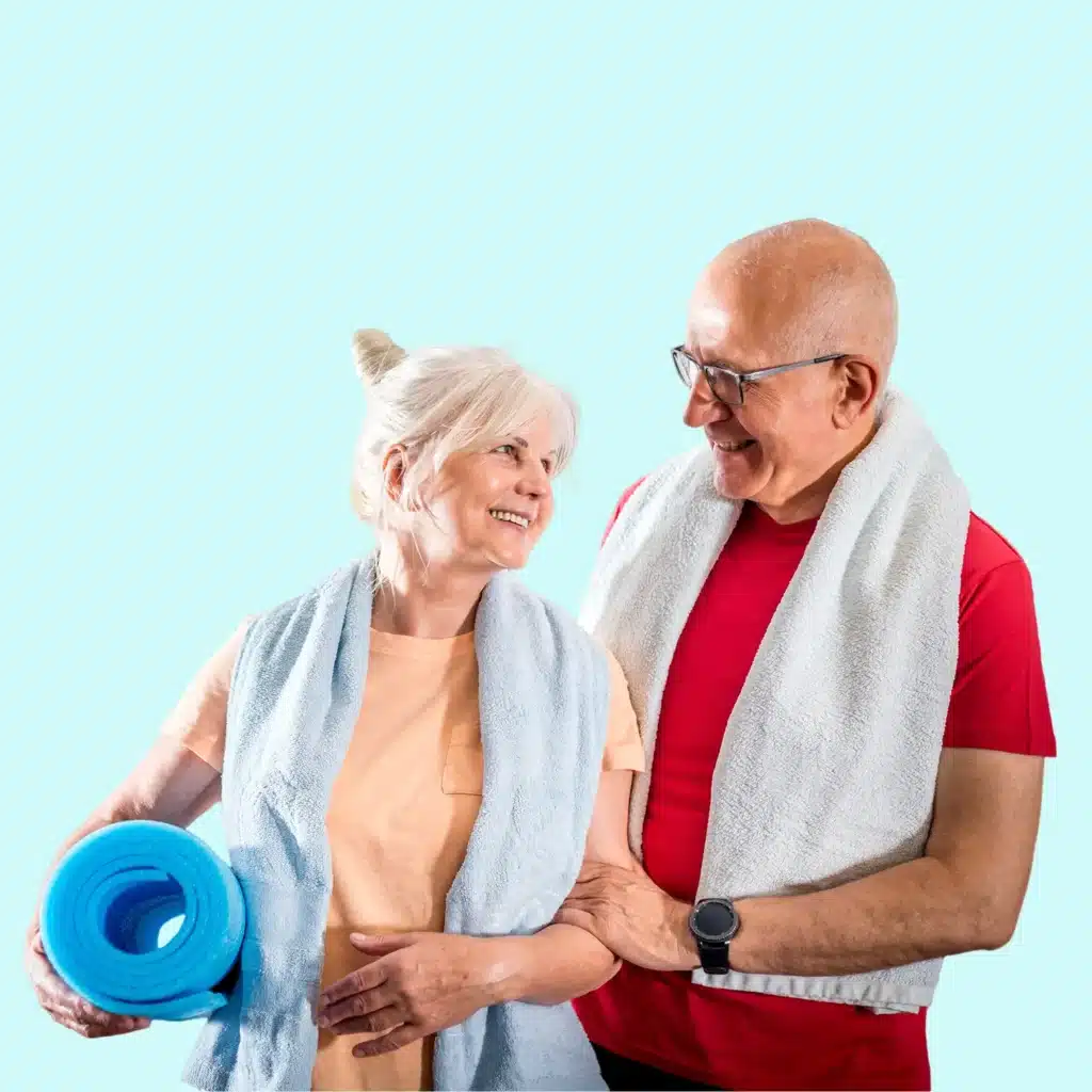 An older woman and man, both smiling, stand together with towels around their necks. The woman holds a blue yoga mat, and the man wears a smartwatch. They appear happy and ready for exercise against a light blue background.