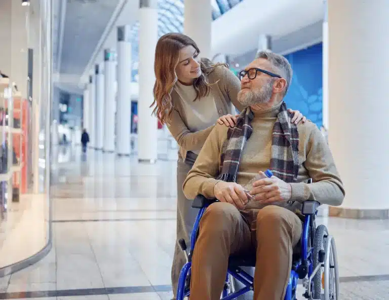 A young woman stands behind an older man in a wheelchair, gently resting her hands on his shoulders. They are indoors in a modern shopping centre, both smiling and sharing a warm, affectionate moment.