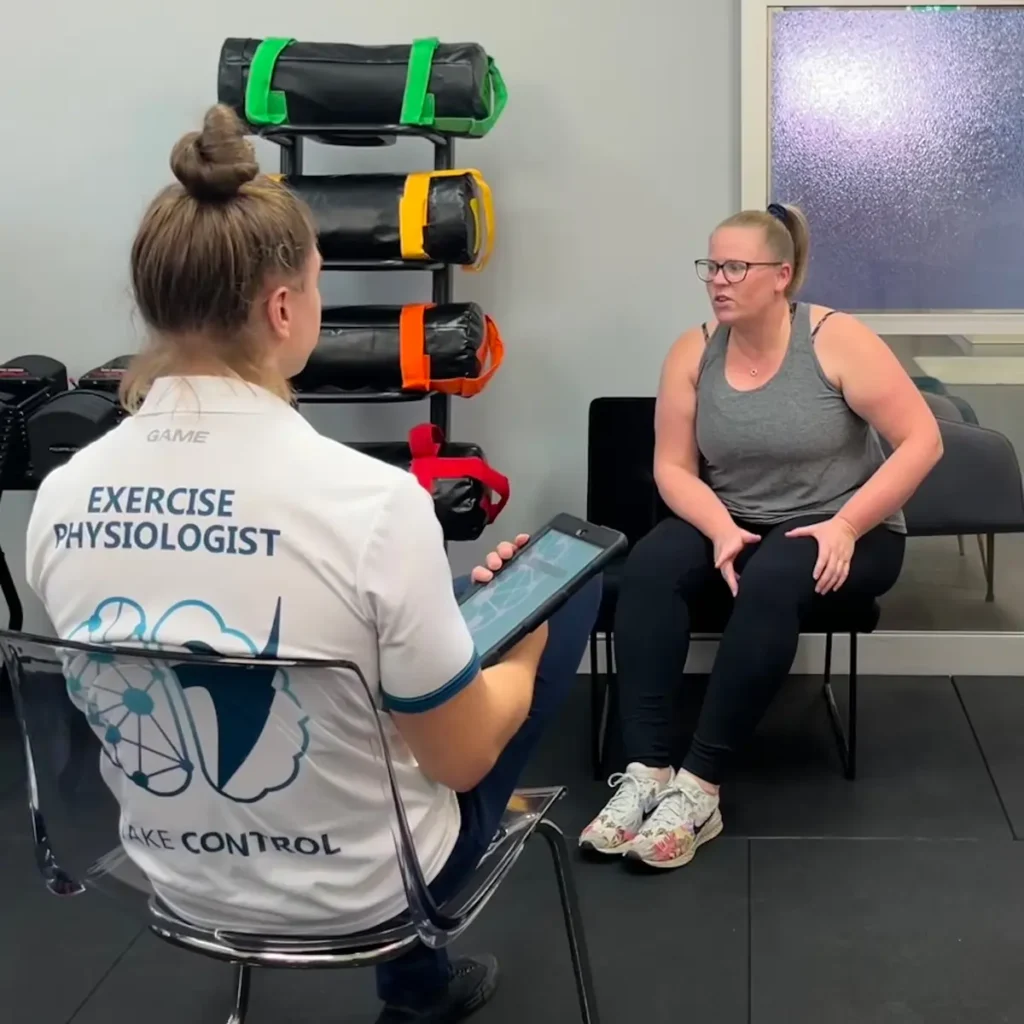 An exercise physiologist with a tablet consults a woman seated in workout clothes in a gym setting, with colourful fitness equipment in the background.