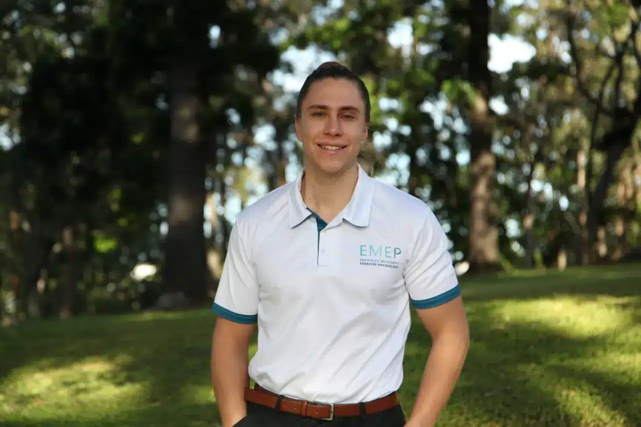 A young man stands outdoors on grass with trees in the background, smiling and wearing a white polo shirt with EMEP and blue accents. His hands are in his pockets.