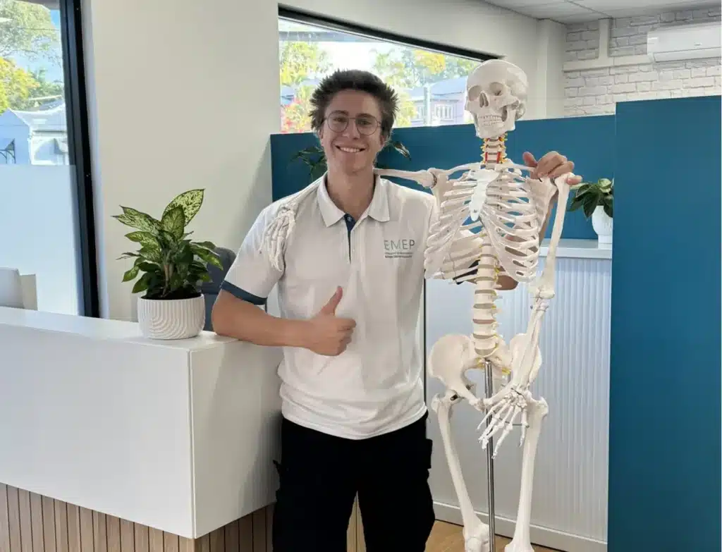 A smiling person in a white polo shirt stands indoors, giving a thumbs-up and posing with a human skeleton model. There are green plants and modern office décor in the background.