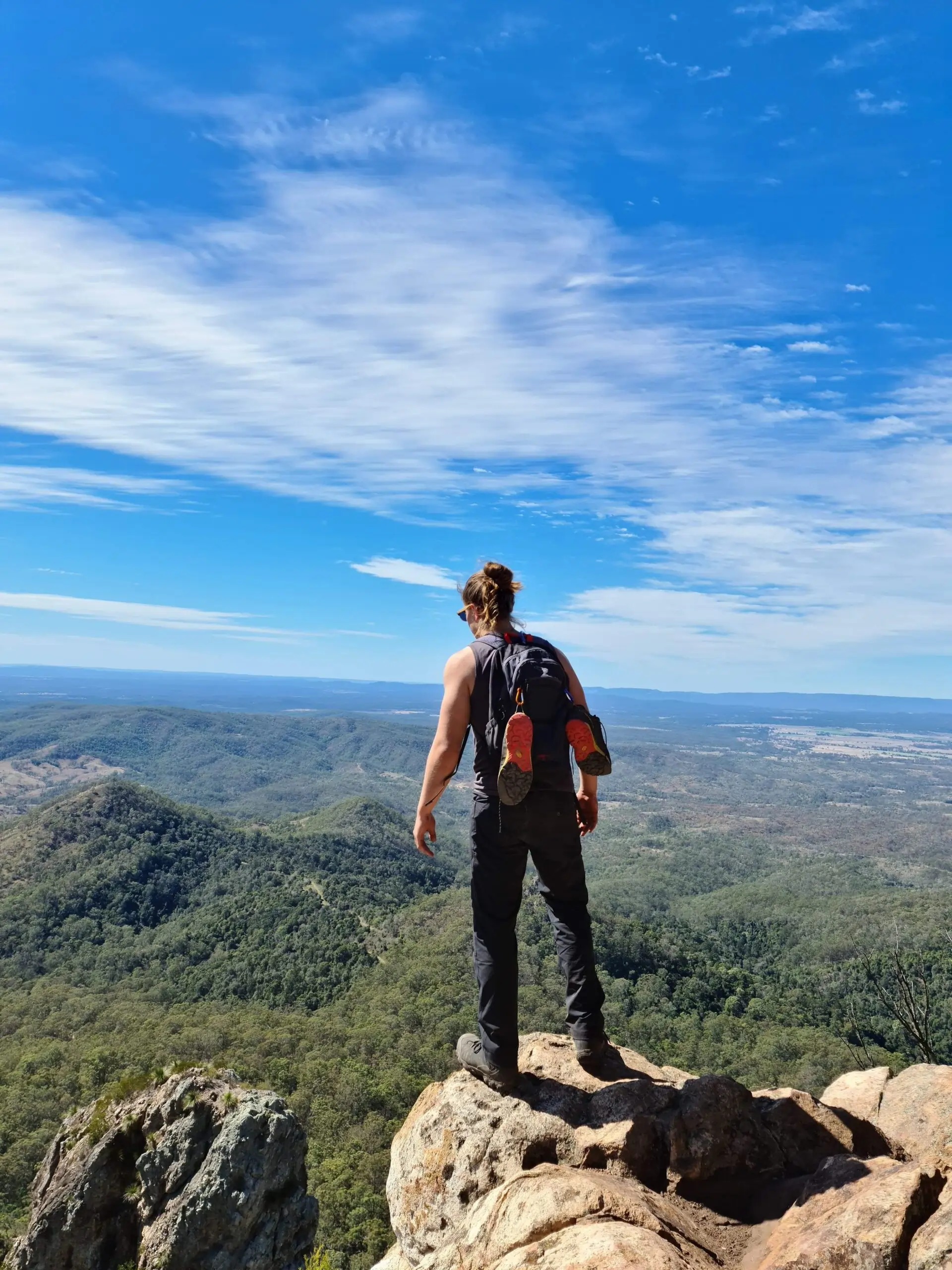 A person with a rucksack stands on a rocky outcrop, overlooking a vast landscape of green hills and forests under a blue sky with scattered clouds.