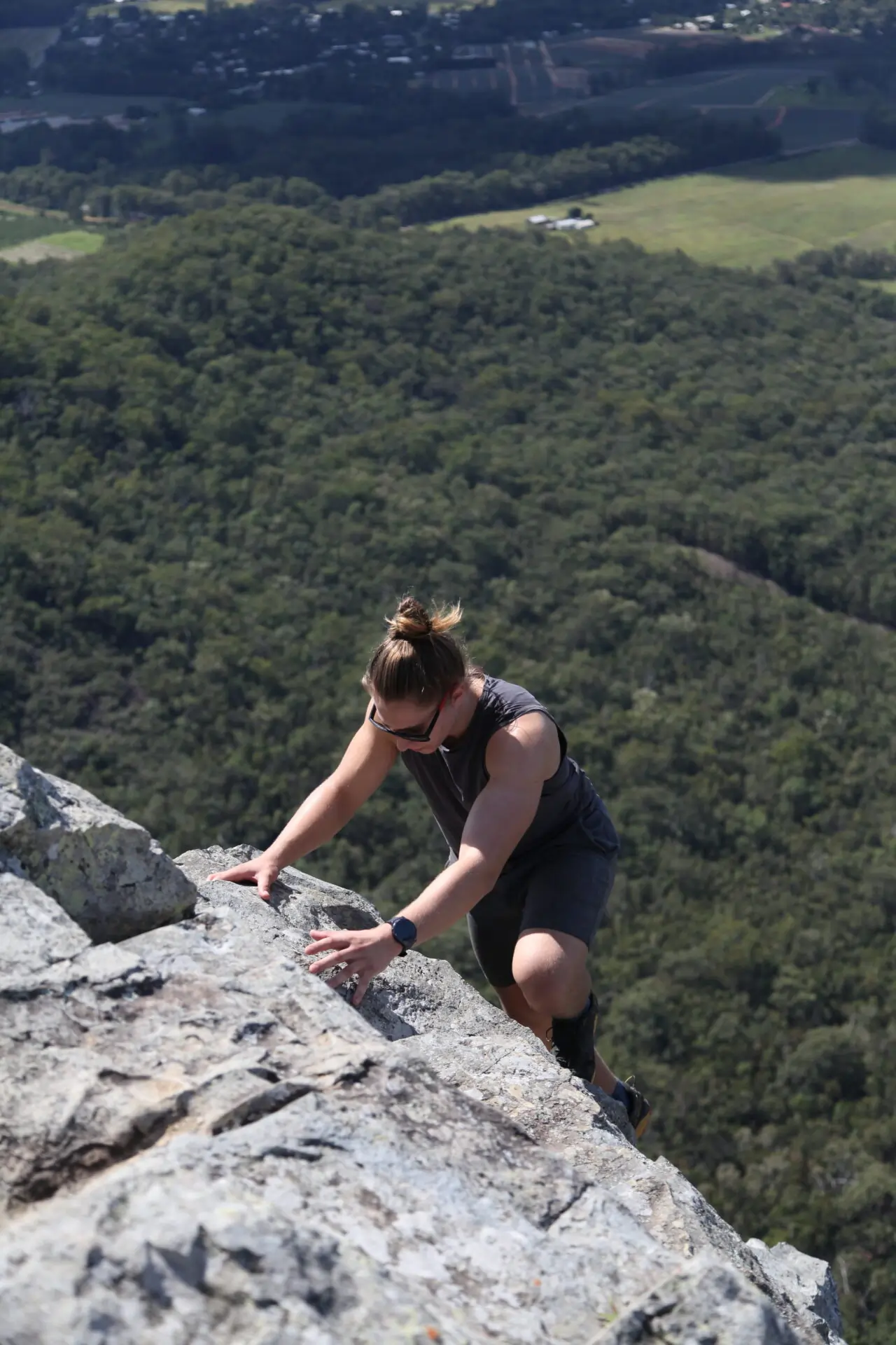 A person wearing sunglasses and a sleeveless vest climbs a steep rocky cliff with a forested valley in the background under bright daylight.