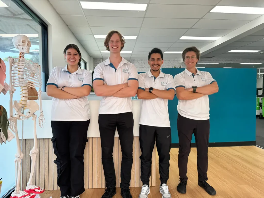 Four people stand indoors in front of a counter, smiling with arms folded. They wear matching white polo shirts and black trousers. A human skeleton model stands to the left. The setting appears to be a clinic or office.