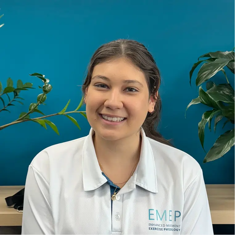A young woman with long brown hair tied back, wearing a white collared shirt with the EMEP logo, smiles at the camera. She is seated in front of a teal wall with green plants on either side.