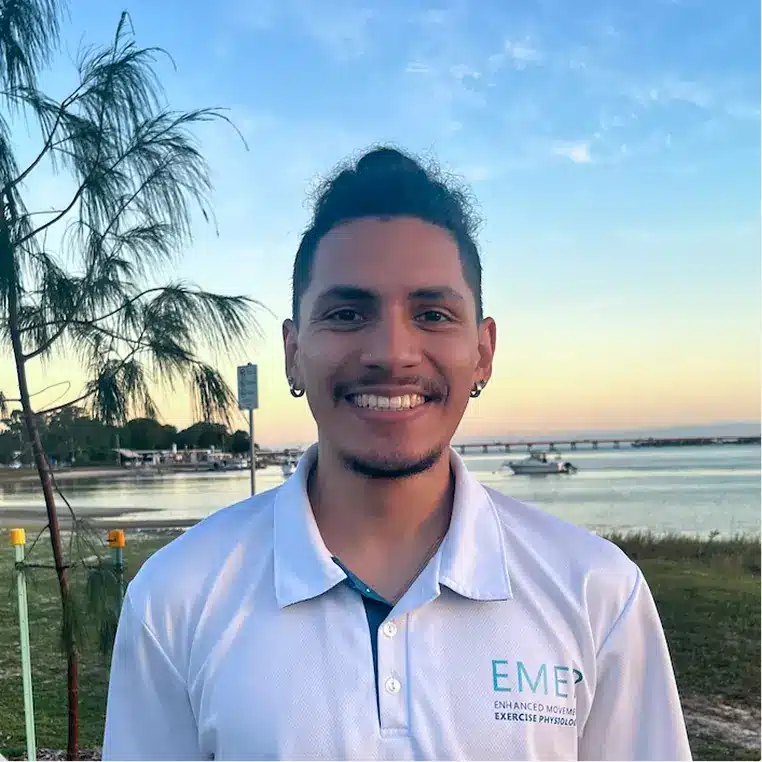 A smiling man with short curly hair and earrings stands outdoors by a body of water, wearing a white EMEP exercise physiology shirt. Trees and a bridge are visible in the background under a blue sky.