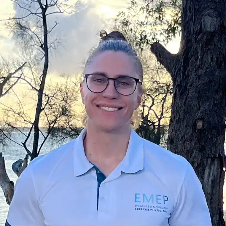 A person with long hair in a bun and glasses smiles outdoors, wearing a white EMEP polo shirt. Trees and water are visible in the background at sunset.