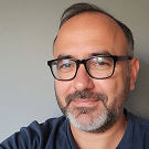 A middle-aged man with short hair, a beard, and glasses is smiling slightly. He is wearing a dark shirt and standing in front of a plain, light-coloured wall.