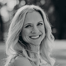 Black and white photo of a smiling woman with long, wavy fair hair, outdoors with a blurred background of trees and sunlight.