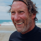 A man with tousled hair and a beard smiles whilst standing on a beach, wearing a black wetsuit. The sea and sky are visible in the background.