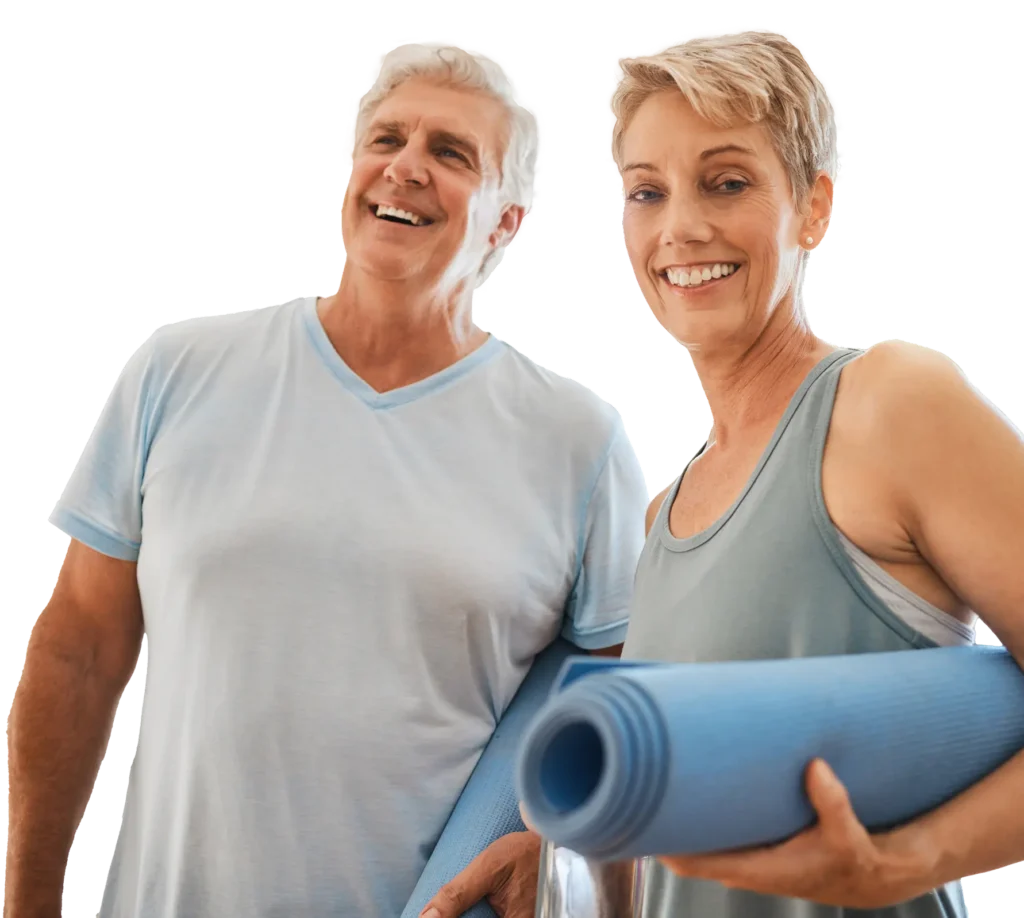 Two older adults, a man and a woman, smiling and holding rolled-up yoga mats, dressed in casual workout clothes, appearing happy and ready for exercise.