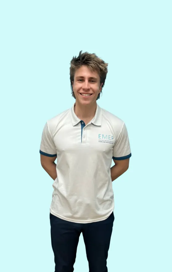 A person with short, spiky brown hair stands smiling, wearing a white collared shirt with blue trim and EMEP text on the chest, and dark trousers, against a light blue background. Hands are positioned behind their back.