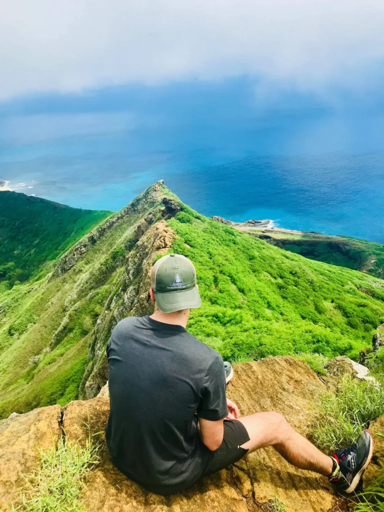 A person wearing a black shirt, black shorts, and a green cap sits on a rocky ledge overlooking a lush green mountain ridge and the blue sea below under a cloudy sky.