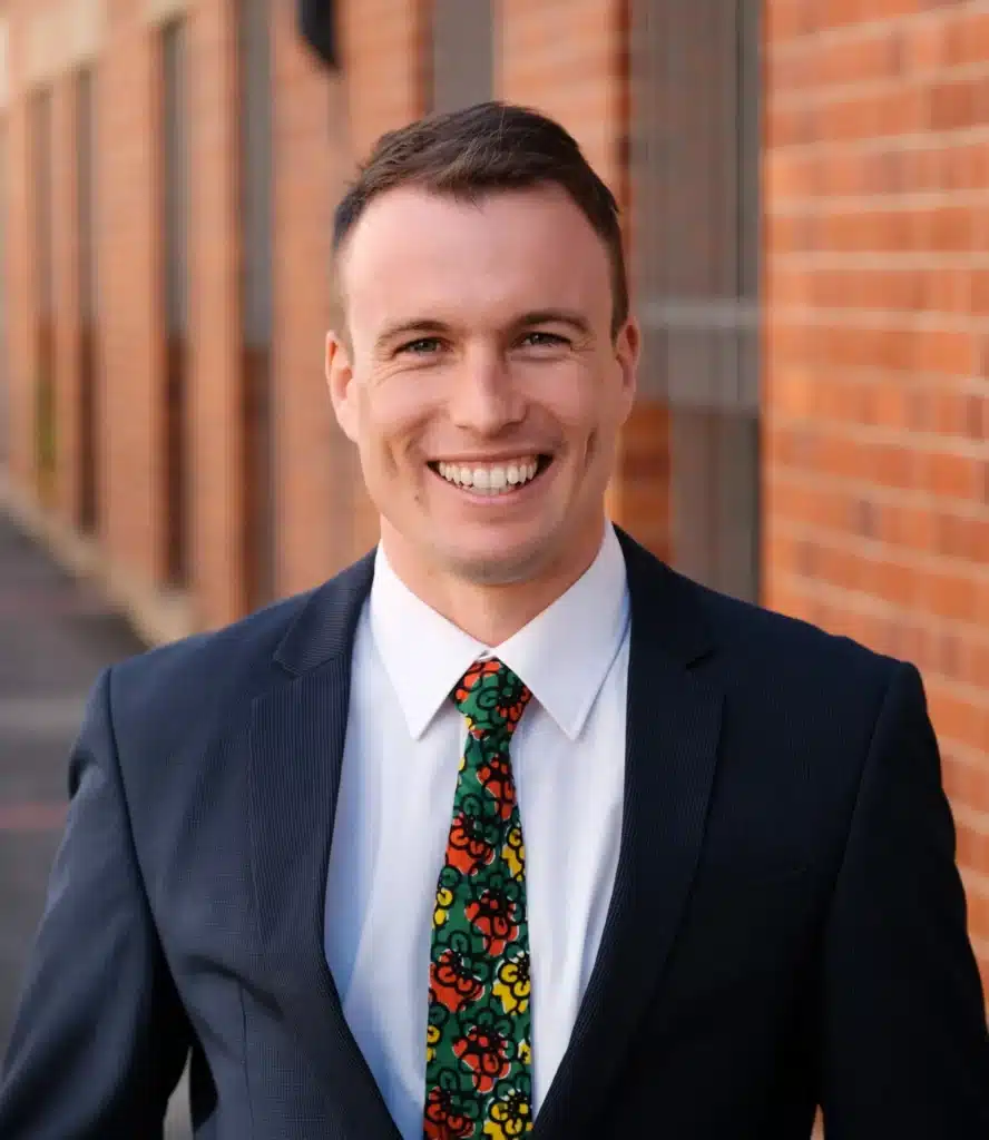 A smiling man in a dark suit, white shirt, and colourful floral tie stands in front of a brick wall, looking directly at the camera.