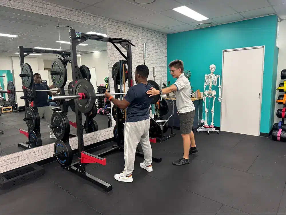 A trainer assists a person preparing to lift a barbell at a gym. Weight plates are racked nearby, and a skeleton model stands in the background against a teal wall.