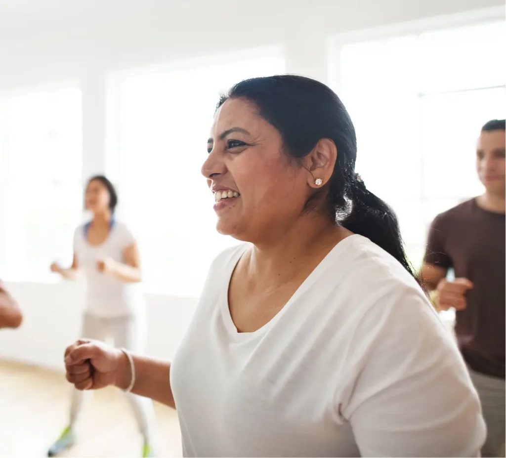 A woman in a white shirt smiles whilst exercising in a bright, sunlit room with other people, all appearing to participate in a group fitness class.