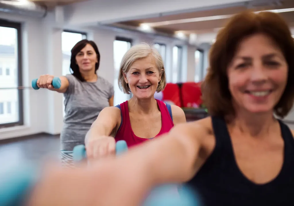 Three women in workout clothes exercise together indoors, each holding a light blue dumbbell and smiling. They are standing in a row, focussing on their fitness routine in a bright room with large windows.
