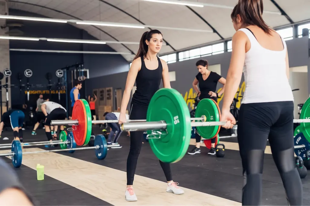 Two women, dressed in black sportswear, face each other while lifting barbells with green and red weight plates in a gym. Other people are exercising in the background.